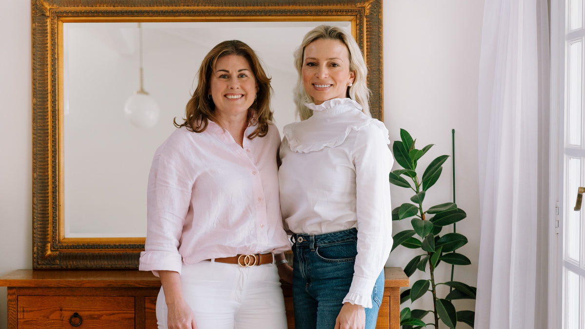 Two women standing side-by-side in front of a large gold-framed mirror on a wooden chest of drawers, with a green plant and white curtains in a bright room.